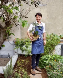 Man wearing a blue apron holding a small potted bonsai tree in an outdoor garden setting with green plants and stone paving.