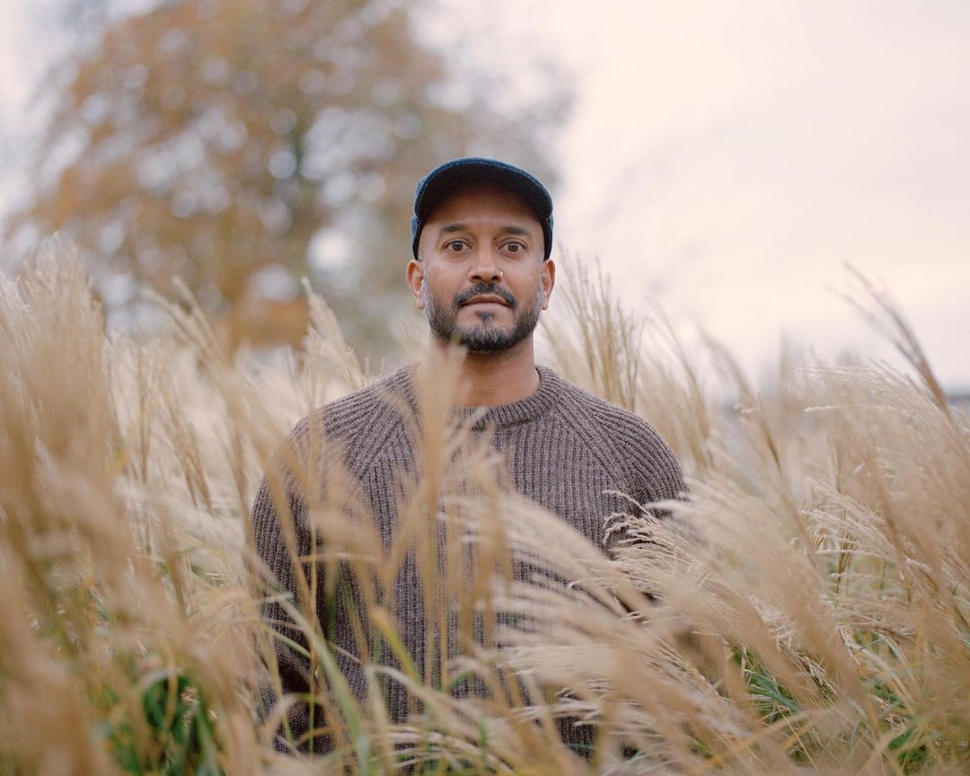 Man in cap stands in a field of tall wheat-colored grass in a medium shot, wearing brown knit sweater on an overcast day.