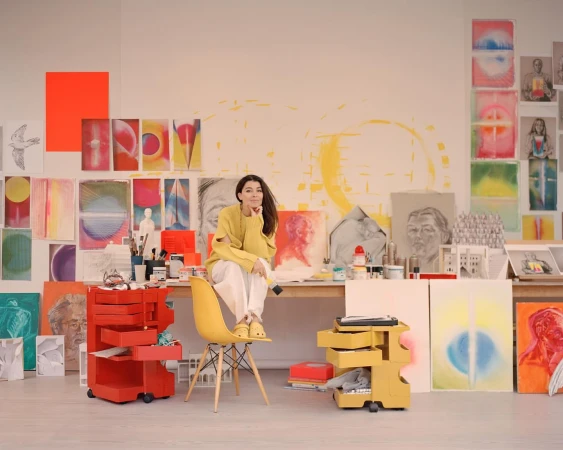 A woman with long brown hair is sitting on a yellow chair in an art studio surrounded by paintings, art supplies, and colorful art carts.