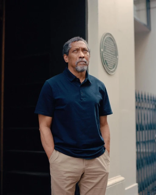 Portrait of a middle-aged African American man in polo shirt and slacks standing outdoors against a building facade with plaque.