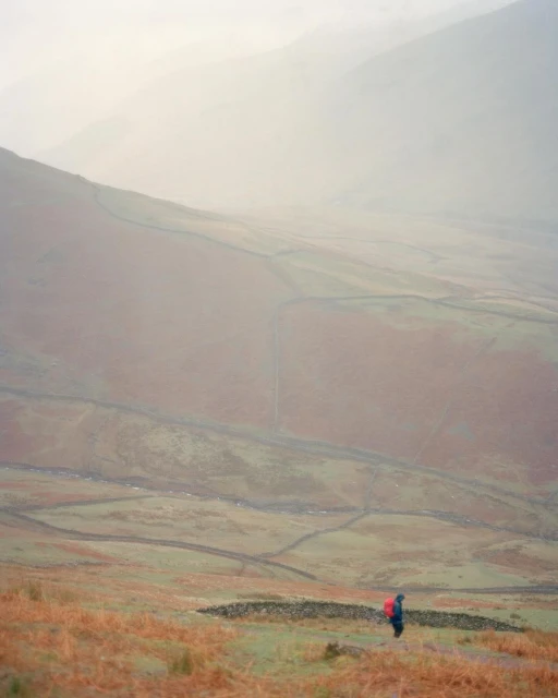 A person with a backpack hikes on a misty, grassy hillside, with rolling hills and mountains in the background under an overcast sky.
