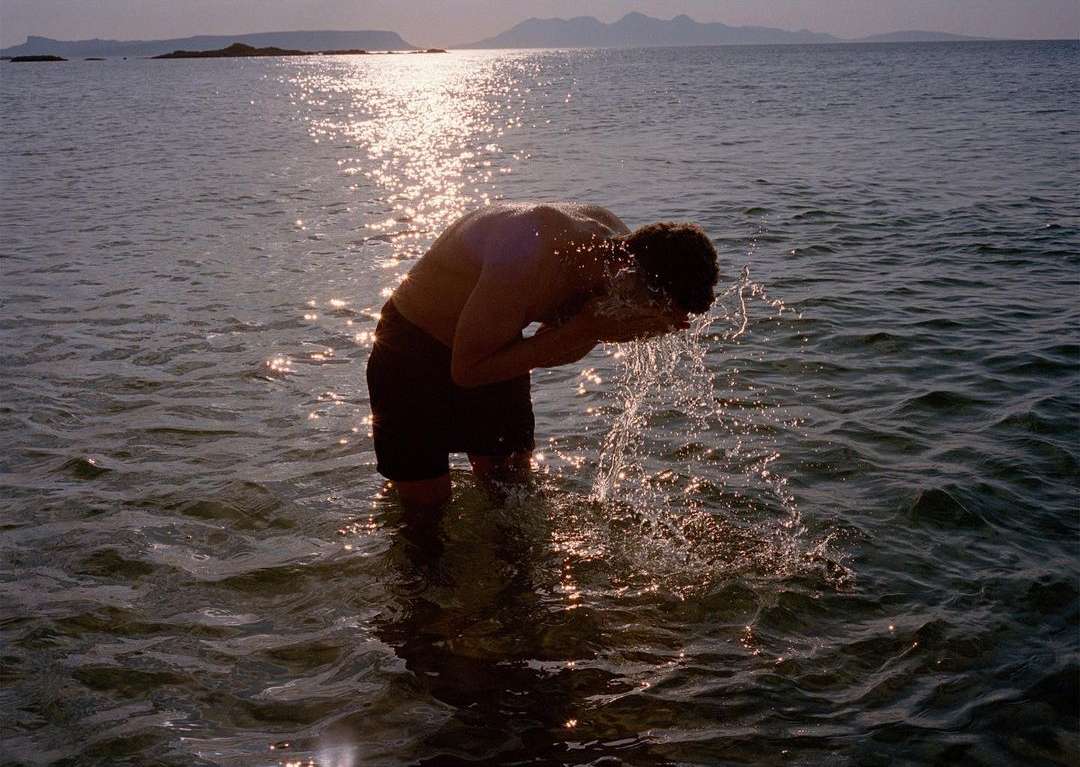 Adult male washing face in shallow ocean water at sunset, with mountains in the background, creating a tranquil scene.