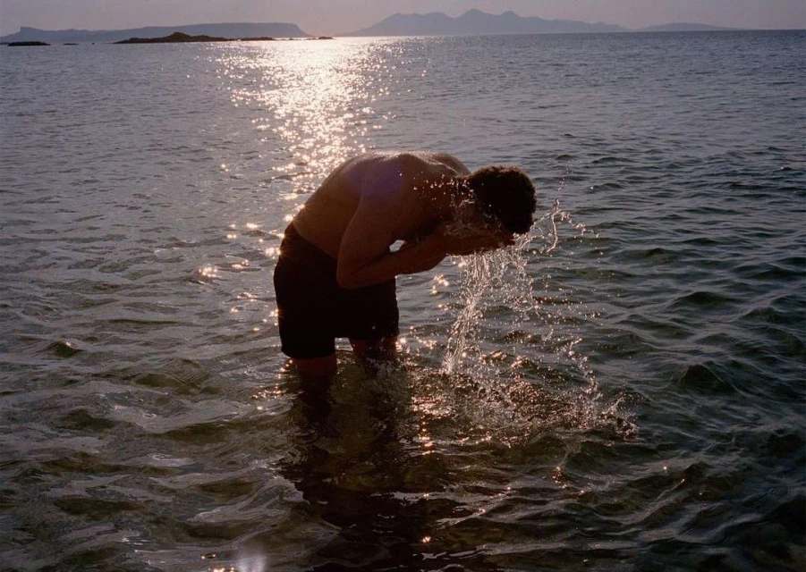 Adult male washing face in shallow ocean water at sunset, with mountains in the background, creating a tranquil scene.