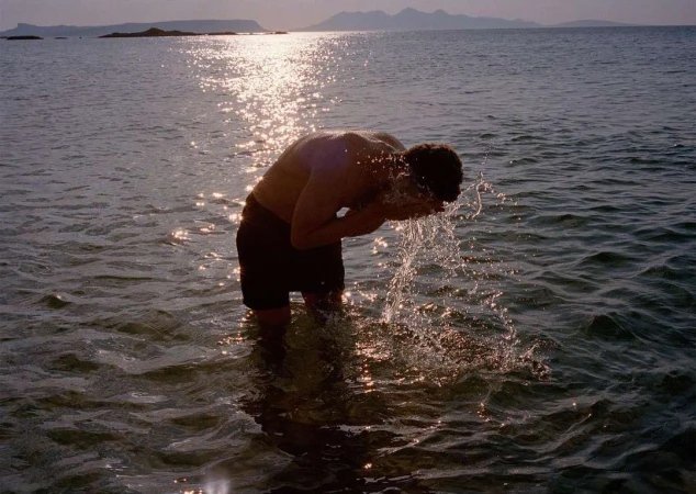 Adult male washing face in shallow ocean water at sunset, with mountains in the background, creating a tranquil scene.