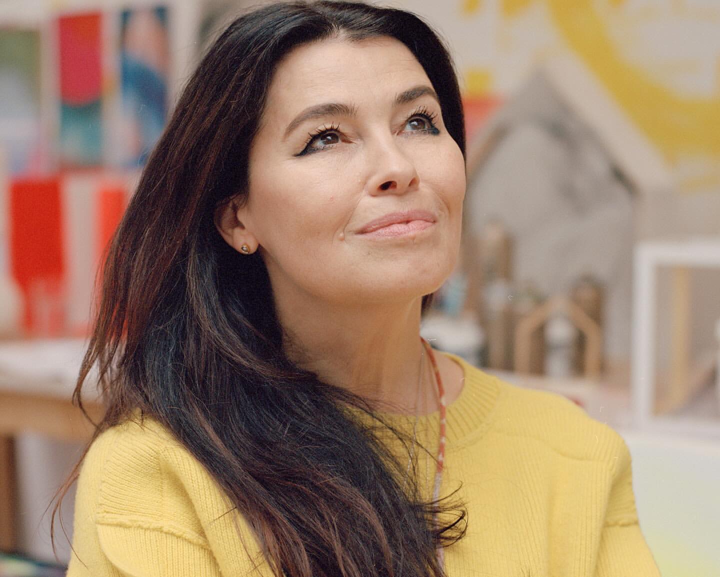 Portrait of an adult woman with long dark hair wearing a yellow sweater, looking up in a brightly lit indoor art studio.