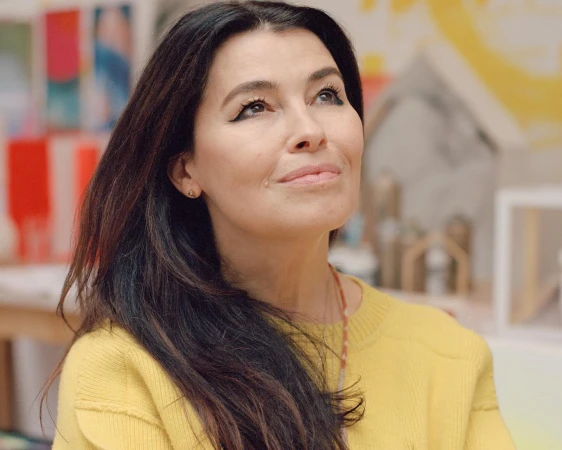 Portrait of an adult woman with long dark hair wearing a yellow sweater, looking up in a brightly lit indoor art studio.