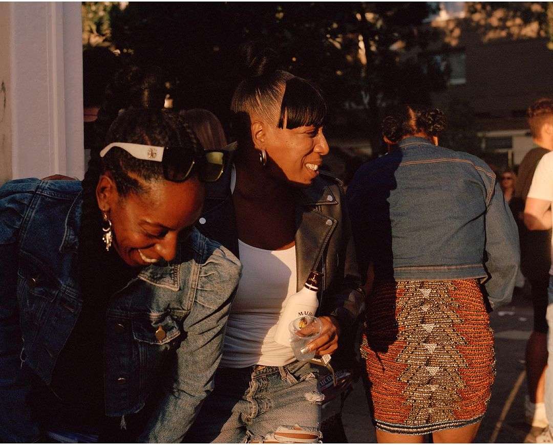 Three adult women enjoying an outdoor gathering; two are smiling, one is seen from the back wearing a decorative skirt, in natural light.