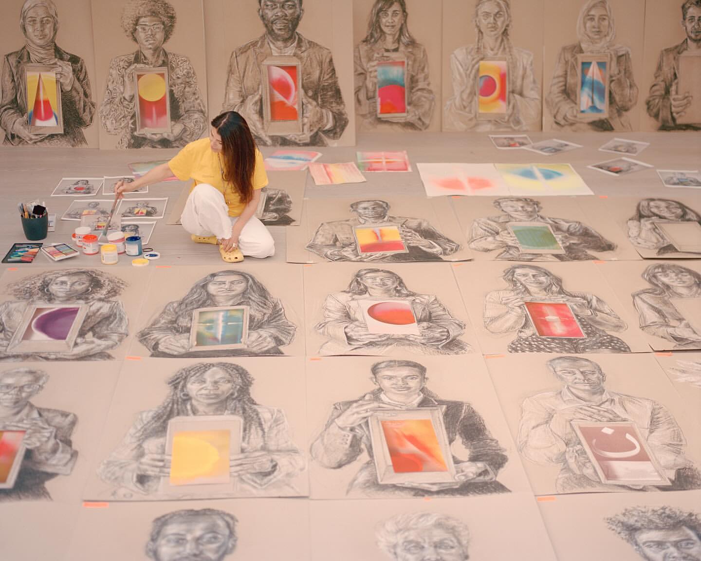 Artist surrounded by charcoal portrait sketches holding painted rectangles, creating a colorful display in a bright, creative studio space.