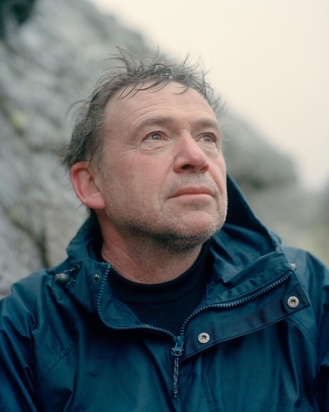 Close-up portrait of an adult man wearing a blue jacket looking up against a blurred rock background on an overcast day.