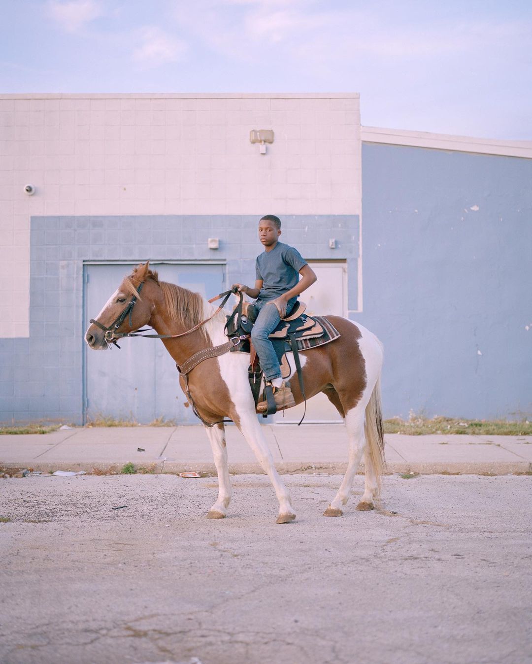 Young man in casual clothes sits astride a paint horse on an asphalt surface with a pale blue and white building in the background.