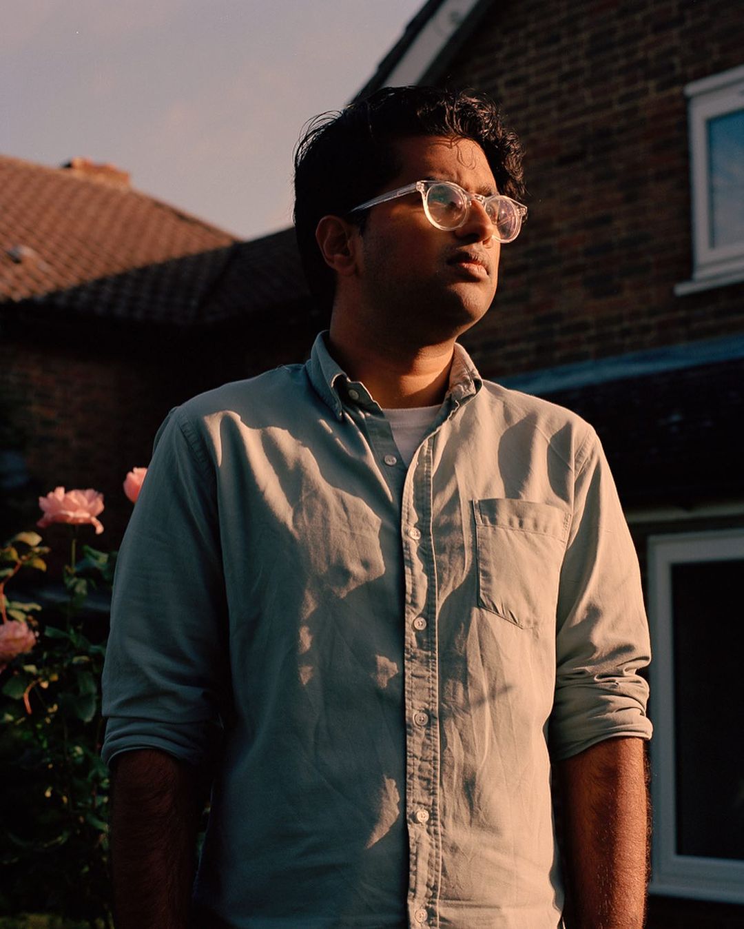 Portrait of a young man with glasses in a backyard, wearing a button-down shirt, with a house and roses in the background.