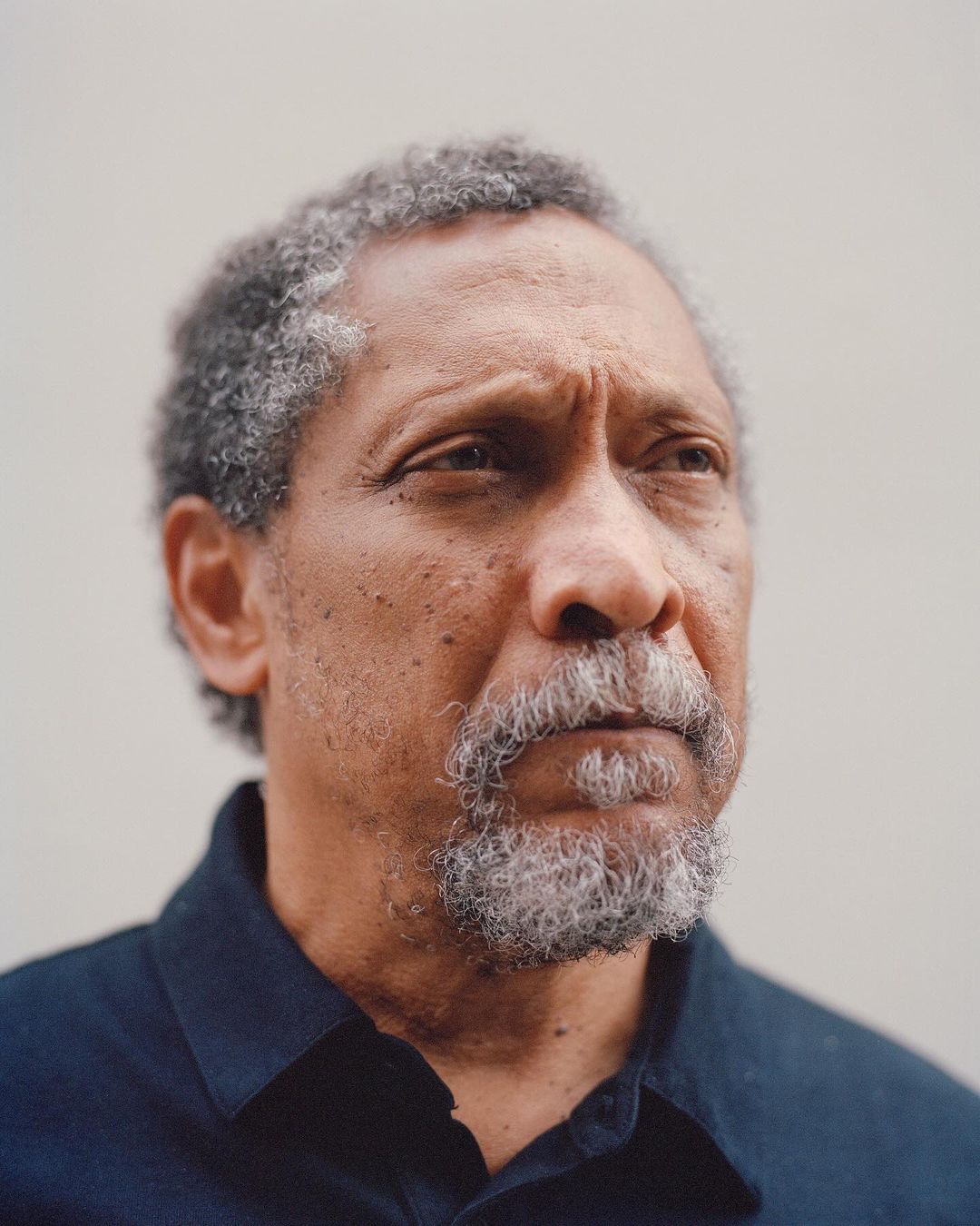 Portrait of a mature adult Black man with grey hair, looking off-camera, wearing a dark button-down shirt against a light background.