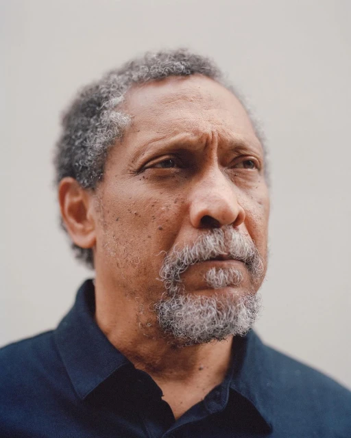 Portrait of a mature adult Black man with grey hair, looking off-camera, wearing a dark button-down shirt against a light background.