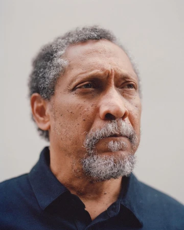 Portrait of a mature adult Black man with grey hair, looking off-camera, wearing a dark button-down shirt against a light background.