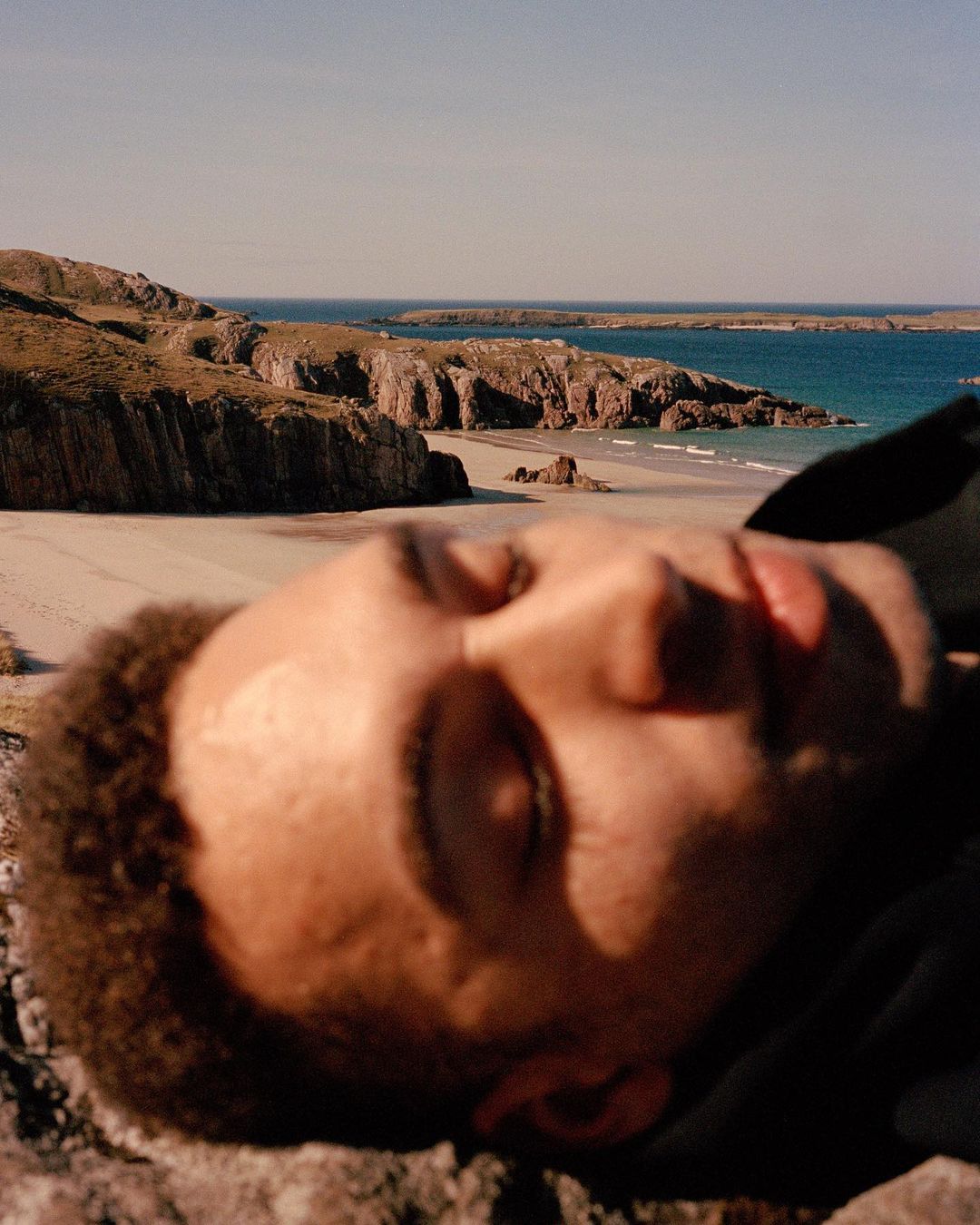 Close-up of a Black male lying on a rock formation with a coastal landscape of cliffs, sandy beach, and turquoise water visible in the background.