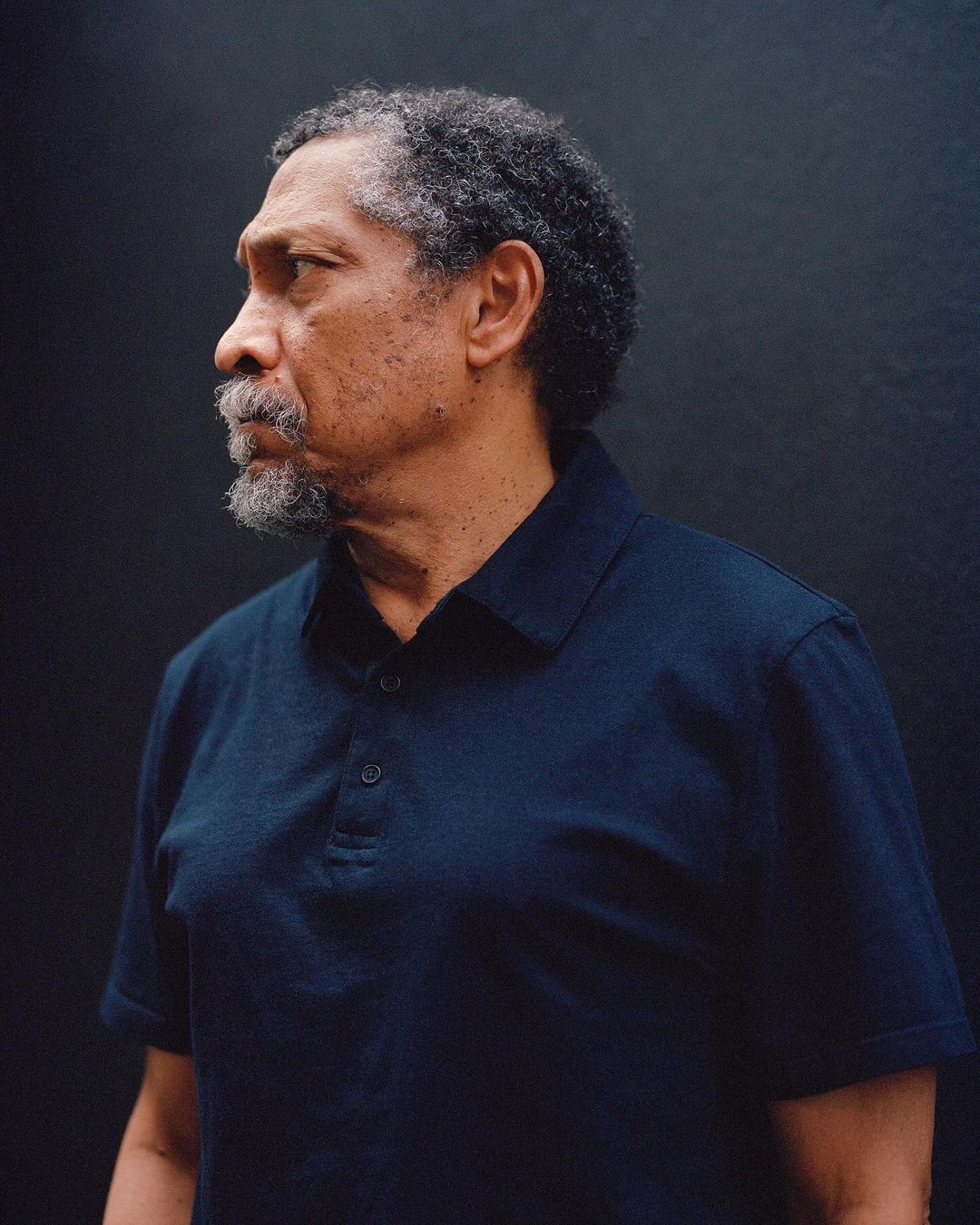 Side-profile portrait of an adult male with salt-and-pepper hair and a beard wearing a blue polo shirt against a dark background.