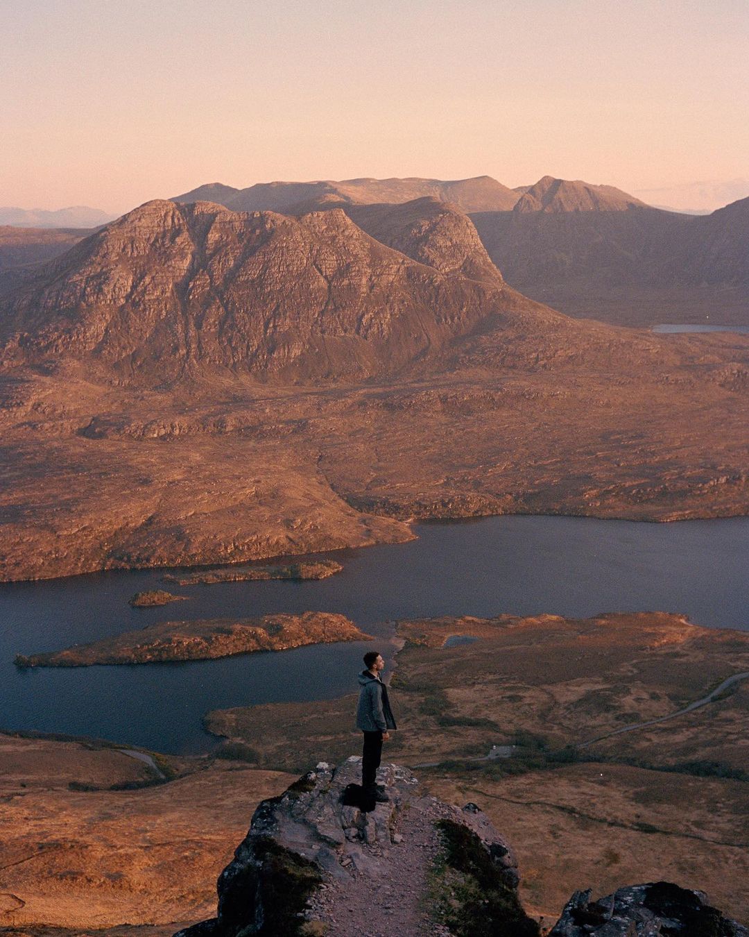 A lone adult stands atop a rocky peak, overlooking a vast landscape of mountains, islands, and a tranquil lake under a soft, muted sky.