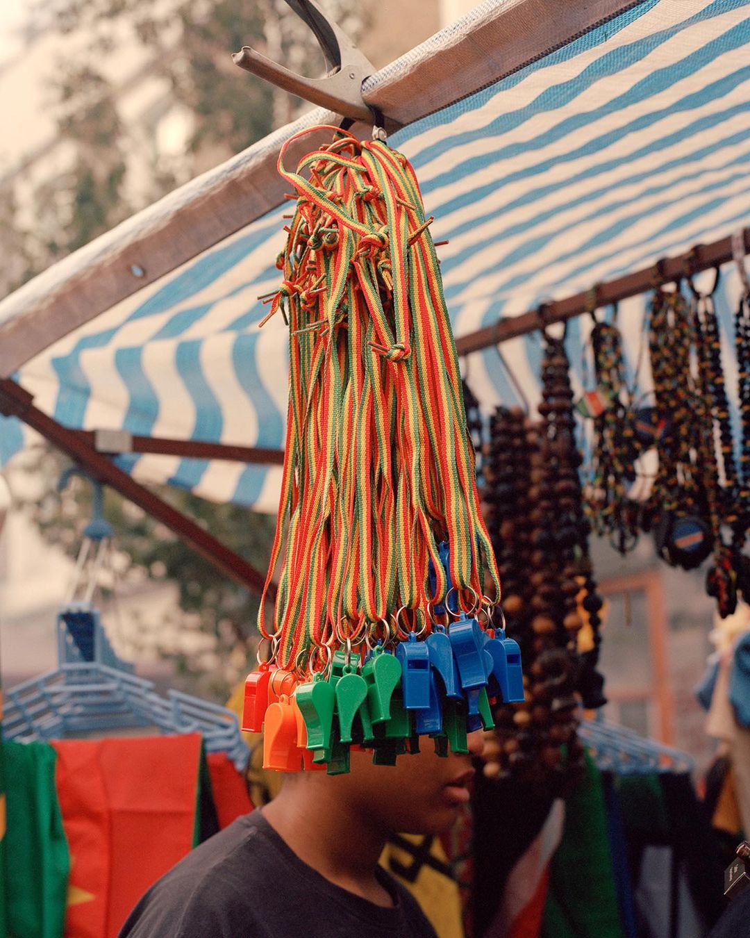 Colorful whistles hanging at an outdoor market under a striped awning, with a person partially visible in the background.