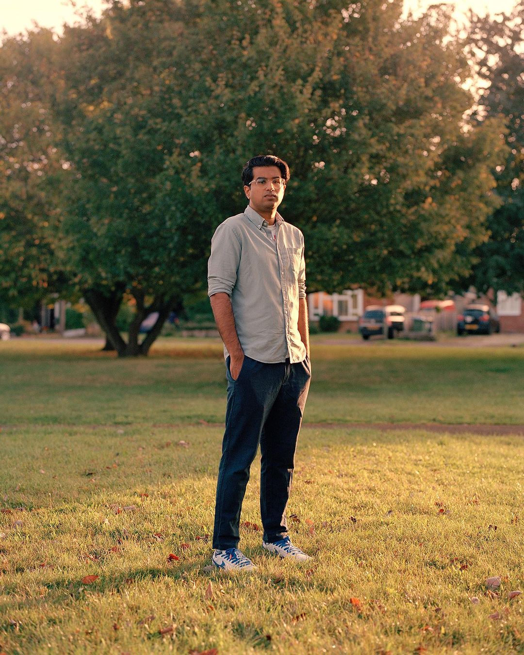 Full body outdoor portrait of a young adult male standing on green grass near large tree in daylight. He wears casual attire.