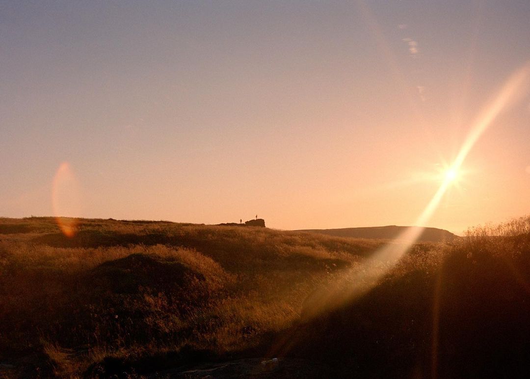 Golden hour landscape with sunlight on grassy hills and two distant people on a hilltop silhouetted against the sky.