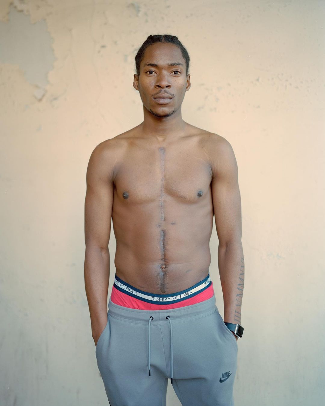 Portrait of a fit, shirtless adult male with braided hair, wearing grey sweatpants, in a direct gaze, against a textured wall.