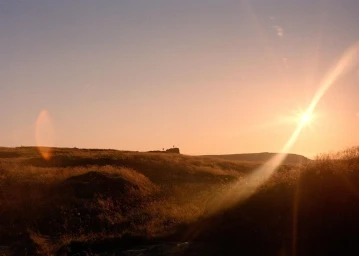 Distant figures silhouetted on a grassy hill during a bright sunset with a prominent lens flare across the sky.