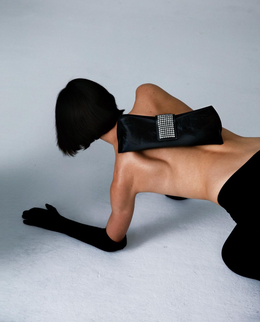 Fashion shot of a slim model with short black hair on all fours wearing a black top, pants, gloves, and clutch purse in studio.