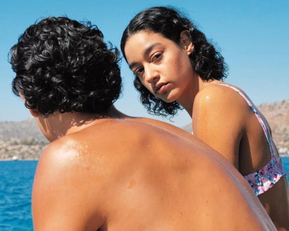 Man and woman with dark curly hair outdoors near bright blue water under a clear sky.