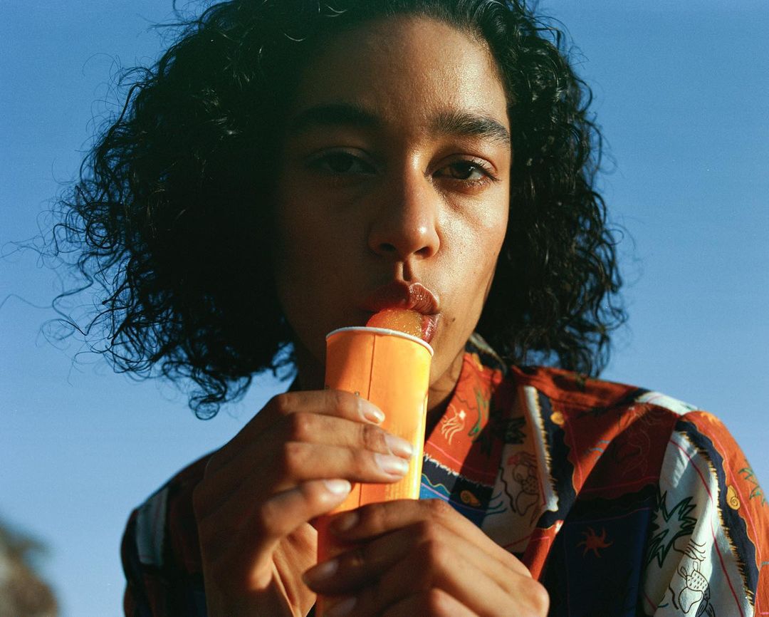 A close-up captures a stylish woman with dark curly hair enjoying a popsicle against a bright blue sky, showcasing a vibrant, summery scene.