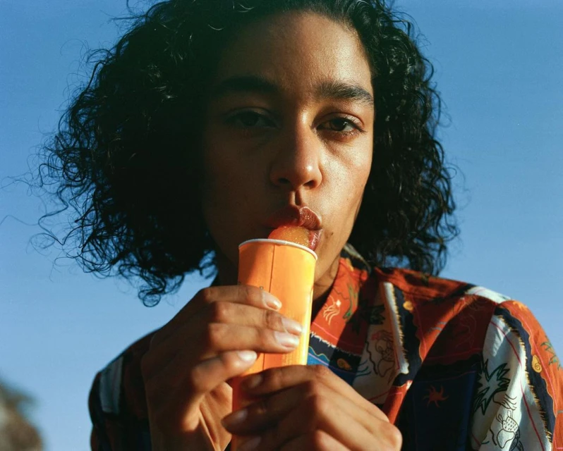 A close-up captures a stylish woman with dark curly hair enjoying a popsicle against a bright blue sky, showcasing a vibrant, summery scene.
