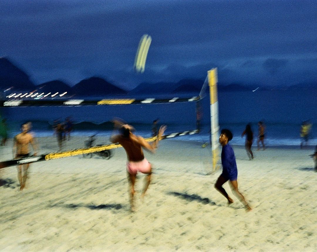 Beach volleyball game at dusk with two shirtless players and others in the background near the ocean and mountains.