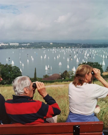 An elderly couple sitting on a bench outdoors looking through binoculars at a large lake filled with white sailboats in Istanbul.