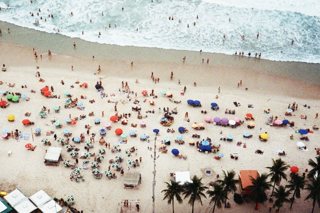 Aerial view of a crowded beach with colourful umbrellas, palm trees, and ocean waves. People are swimming and sunbathing on the sand.
