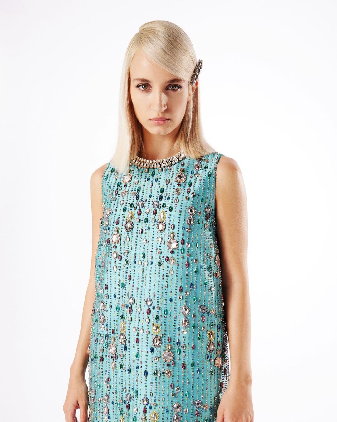A fair-skinned female model with platinum blonde hair poses in a sleeveless, light-blue shift dress adorned with rhinestones against a white backdrop.