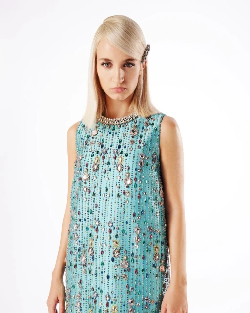 A fair-skinned female model with platinum blonde hair poses in a sleeveless, light-blue shift dress adorned with rhinestones against a white backdrop.