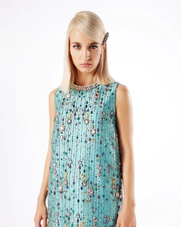 A fair-skinned female model with platinum blonde hair poses in a sleeveless, light-blue shift dress adorned with rhinestones against a white backdrop.