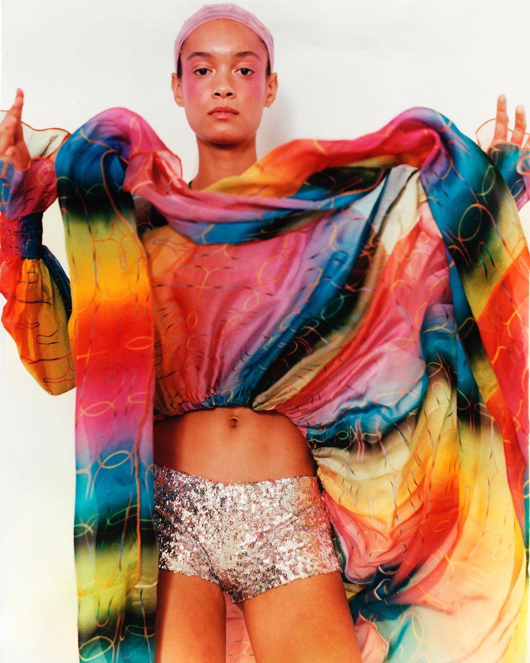 Woman poses in a colorful, flowing top and sequined shorts against a bright backdrop, fashion photography, studio shot.
