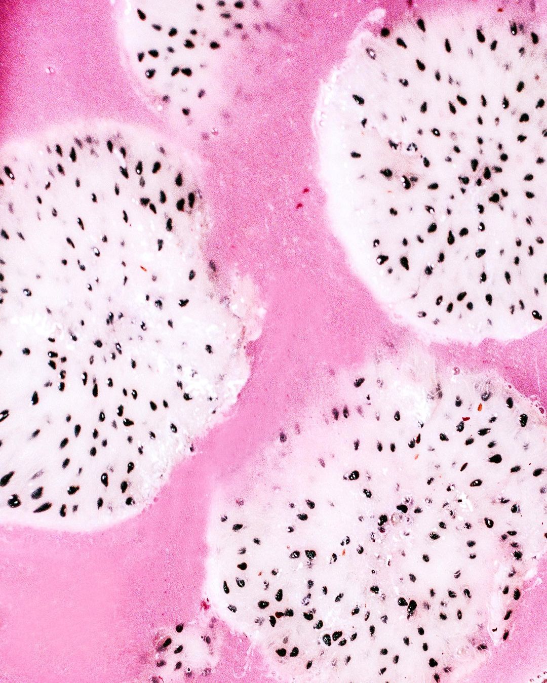 Overhead close-up of sliced dragon fruit with white flesh and black seeds in a pastel pink liquid or gel-like substance, studio lit.