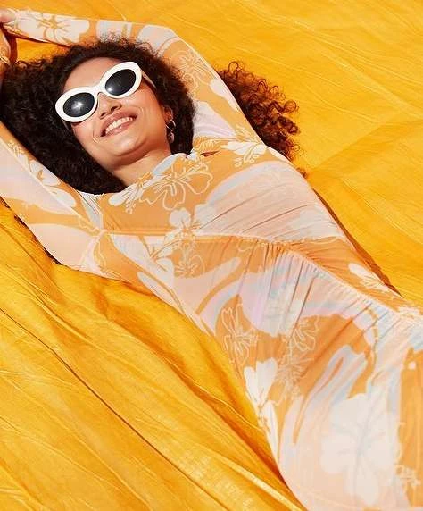A woman with curly hair and white sunglasses lies on a yellow surface, smiling and wearing a patterned dress, looking at the camera.