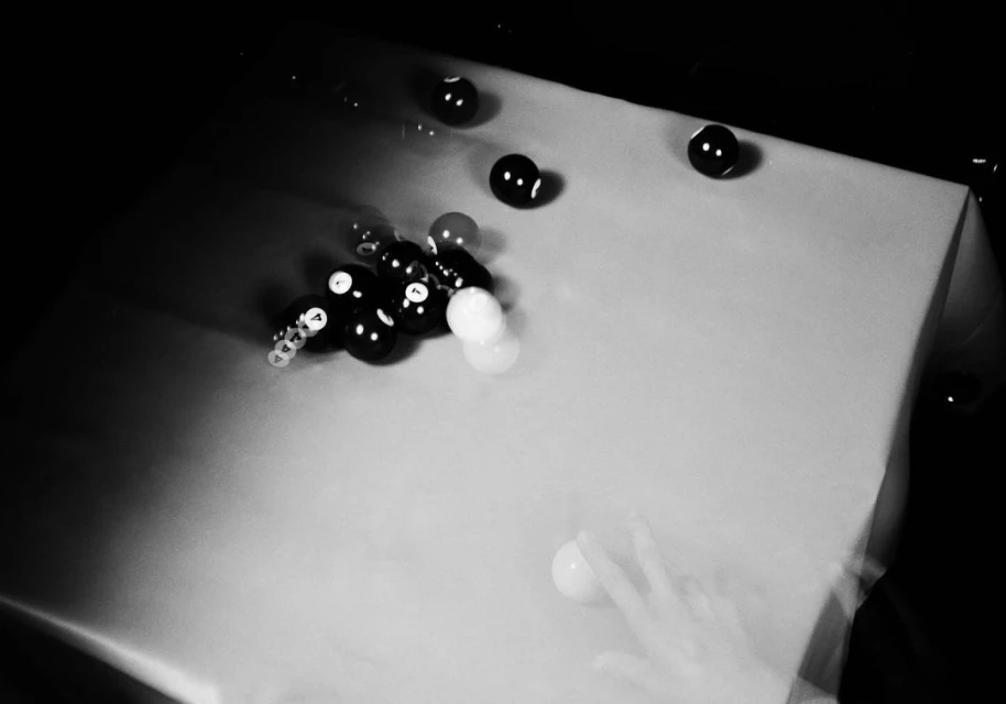 Monochrome shot of a pool table featuring a rack of pool balls, scattered balls, and a cue stick in motion captured with long exposure.