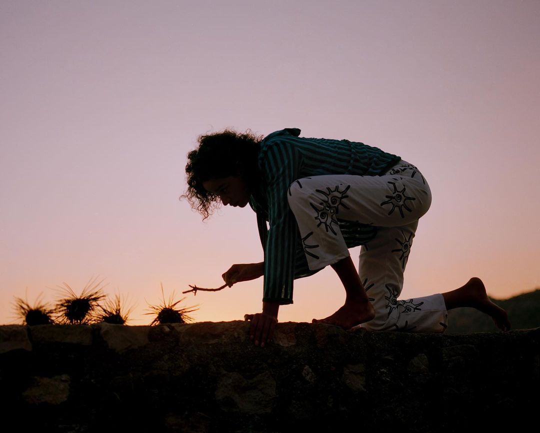 Woman kneels at dusk, interacting with spiky objects on a stone ledge against a soft, colorful sky. Silhouette and barefoot details visible.