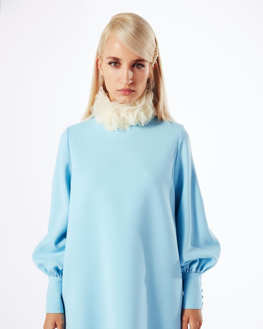 Blonde woman in light blue dress and ruffled collar stands in a bright studio with soft lighting, posing for a fashion editorial portrait.