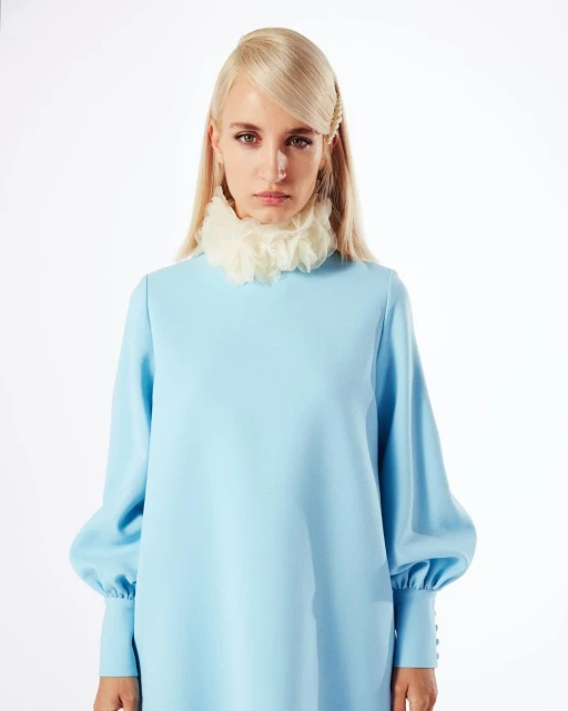Blonde woman in light blue dress and ruffled collar stands in a bright studio with soft lighting, posing for a fashion editorial portrait.