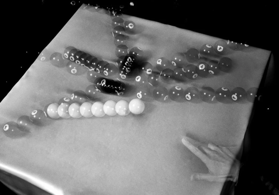Monochrome shot of a pool table with multiple pool balls captured in motion, showing blurred trails and a hand near the edge.