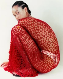 Model with braided hair crouching while wearing a red mesh dress against a neutral white background