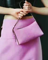 Woman holding a pink suede shoulder bag in front of her waist, wearing a pink skirt and black top against a dark background.