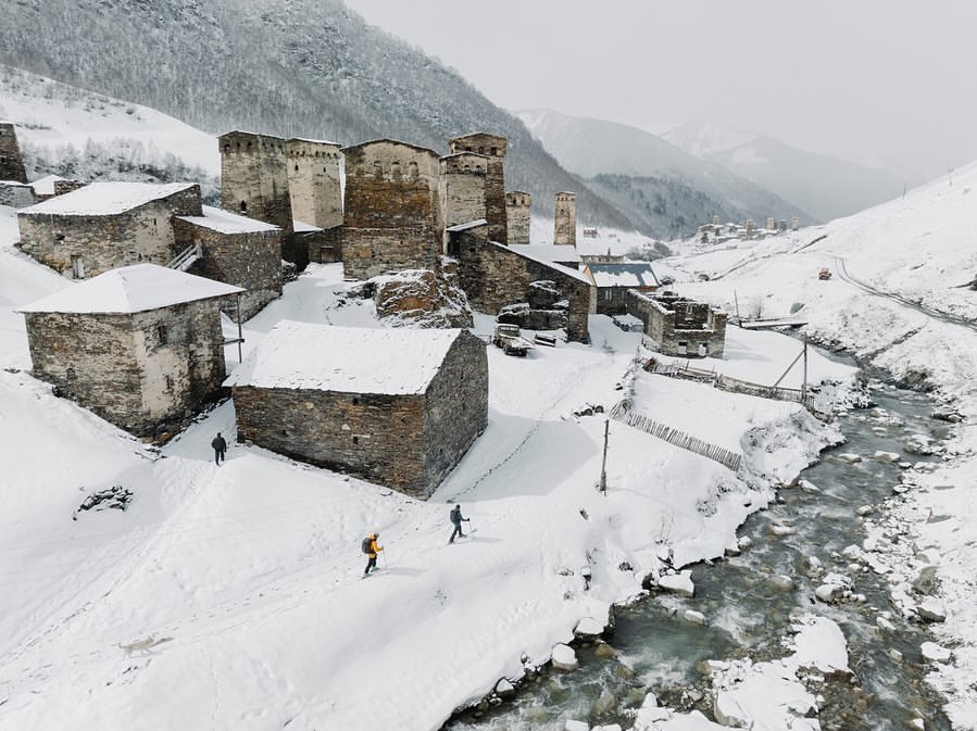 Scenic view of snow-covered stone village nestled amid majestic mountains, with hikers adding scale to the winter landscape.