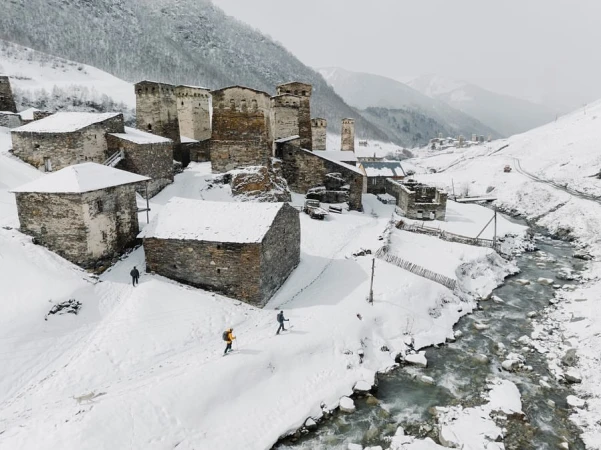 Scenic view of snow-covered stone village nestled amid majestic mountains, with hikers adding scale to the winter landscape.