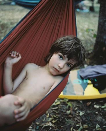 Young child resting inside a red fabric hammock outdoors surrounded by forest floor foliage