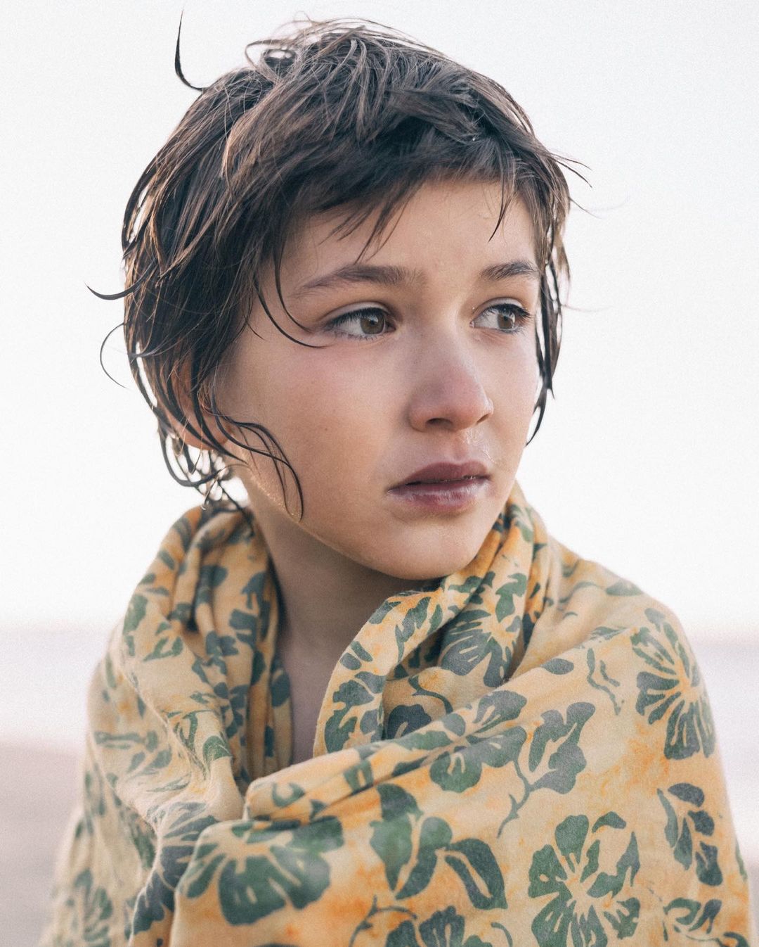Portrait of a young person wrapped in a floral-patterned blanket outdoors, featuring wet hair, brown eyes, and a pensive expression. 